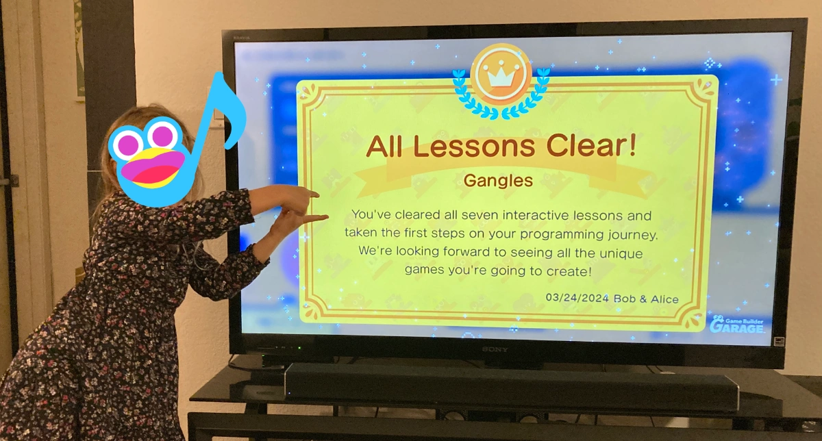 A photo of my daughter posing next to the TV, which shows her completing the final lesson in Game Builder Garage. Her face is obscured with a SFX nodon.